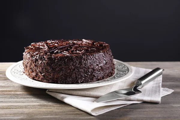 Delicious chocolate cake in plate with napkin and blade on wooden table on dark background — Stock Photo, Image