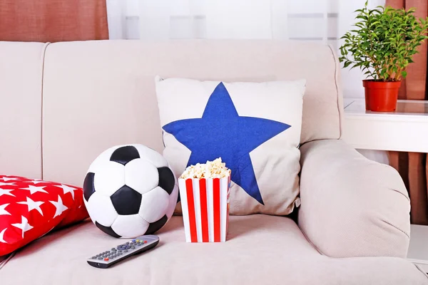 Soccer ball, remote control and box of popcorn on comfortable sofa, indoors — Stock Photo, Image