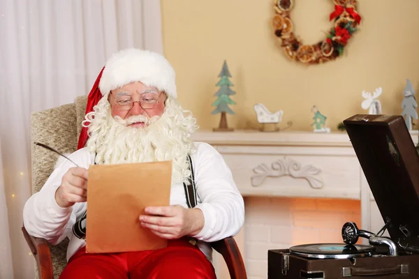 Santa Claus sitting with list of children wishes in comfortable chair near fireplace at home — Stock Photo, Image