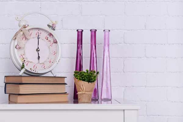 Interior design with alarm clock, plant, decorative vases and stack of books on tabletop on white brick wall background