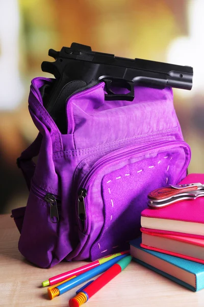 Pistola en la mochila de la escuela en la mesa de madera, sobre fondo brillante —  Fotos de Stock