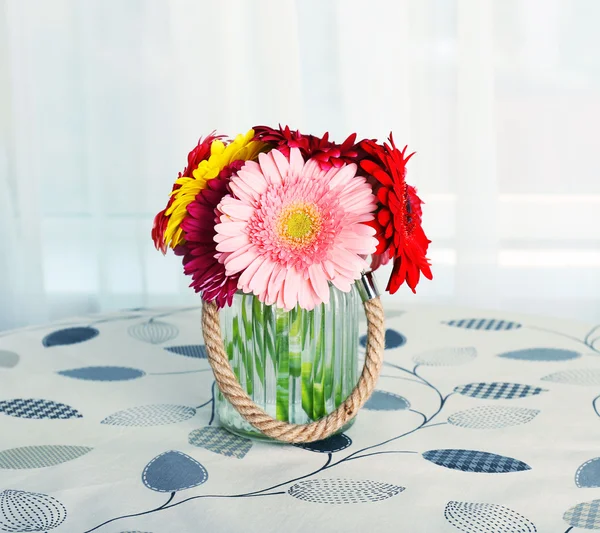 Стеклянная ваза разноцветных цветов герберы на столе на фоне занавесок — стоковое фото