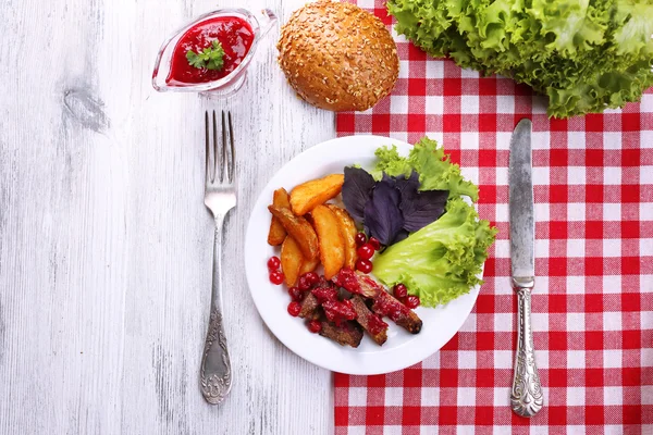 Boeuf à la sauce aux canneberges, tranches de pommes de terre rôties, légumes et pain sur assiette, sur fond de bois couleur — Photo