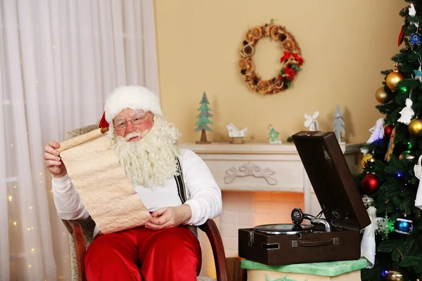 Santa Claus sitting with list of children wishes in comfortable chair near fireplace at home — Stock Photo, Image