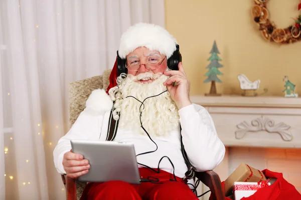 Papai Noel sentado com tablet digital em cadeira confortável perto da lareira em casa — Fotografia de Stock