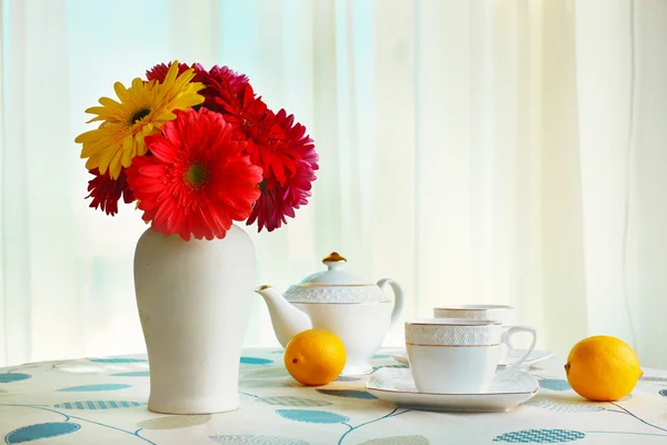 Gerberas coloridas en jarrón con tetera, tazas y limones en la mesa sobre fondo de cortinas —  Fotos de Stock