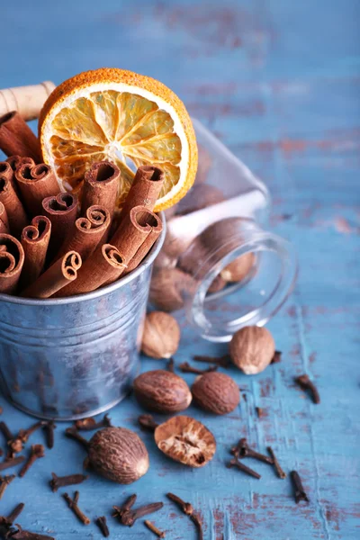 Palitos de canela con naranja seca en cubo de metal, nuez moscada en frasco de vidrio y clavos de olor sobre fondo de mesa de madera de color — Foto de Stock