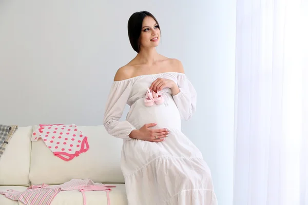 Mulher grávida com botas de bebê sentado no sofá, no quarto — Fotografia de Stock