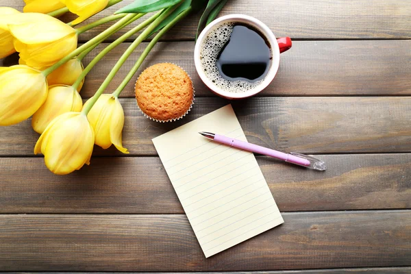 Tasse Kaffee mit frischem Cupcake, Tulpen und leerem Blatt Papier auf Holzgrund — Stockfoto