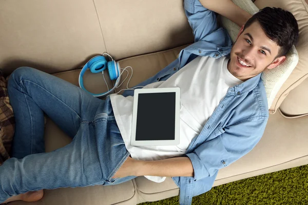 Bonito jovem com tablet deitado no sofá no quarto — Fotografia de Stock