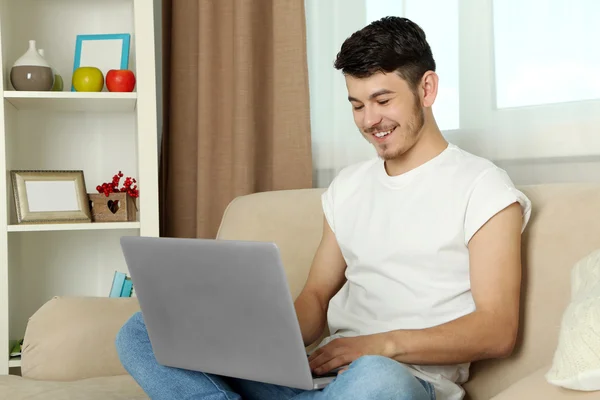 Όμορφος νεαρός άνδρας κάθεται στον καναπέ και χρησιμοποιώντας φορητό υπολογιστή στο δωμάτιο — Φωτογραφία Αρχείου