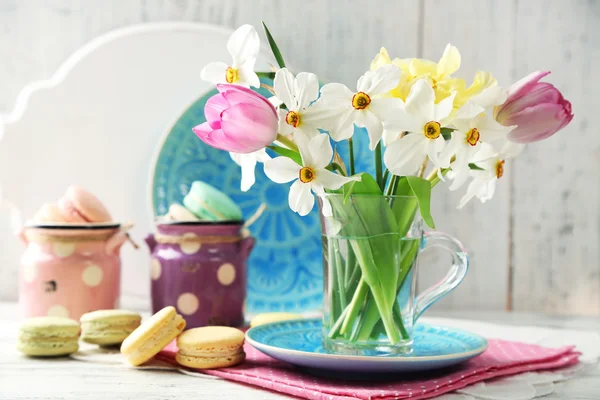 Spring bouquet in glass mug  and tasty macaroons on color wooden background — Stock Photo, Image