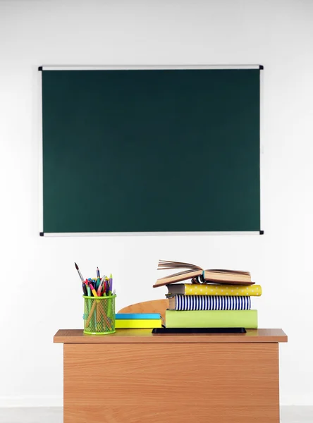 Wooden desk with stationery and chair in class on blackboard background — Stock Photo, Image