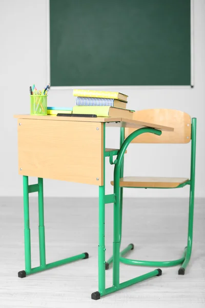 Wooden desk with stationery and chair in class on blackboard background — Stock Photo, Image