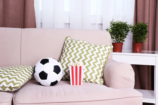 Soccer ball and box of popcorn on comfortable sofa, indoors — Stock Photo, Image