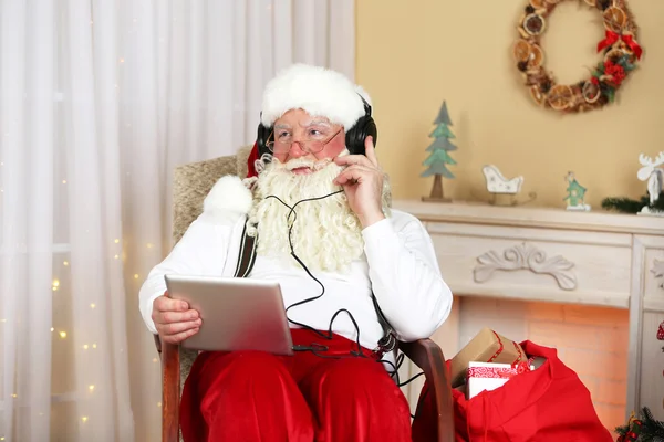 Santa Claus sentado con tableta digital en silla cómoda cerca de la chimenea en casa — Foto de Stock