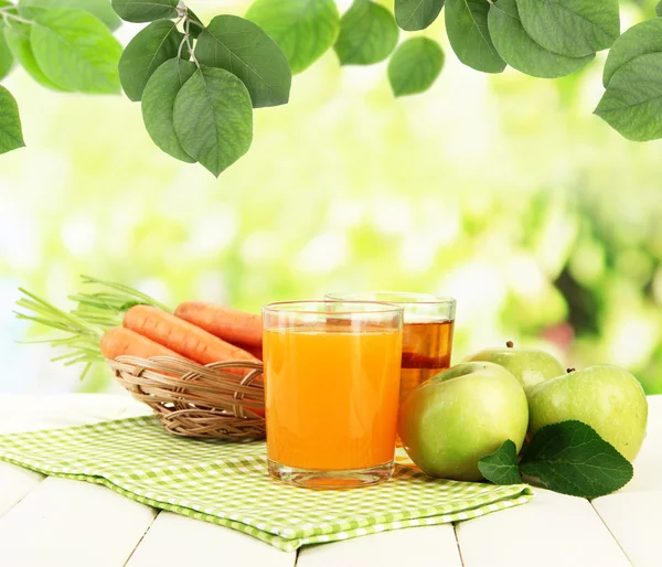 Glazen van SAP, appelen en wortelen op tafel op natuurlijke achtergrond — Stockfoto