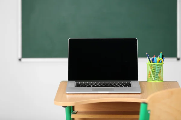 Mesa de madeira com artigos de papelaria e laptop em sala de aula sobre fundo quadro-negro — Fotografia de Stock