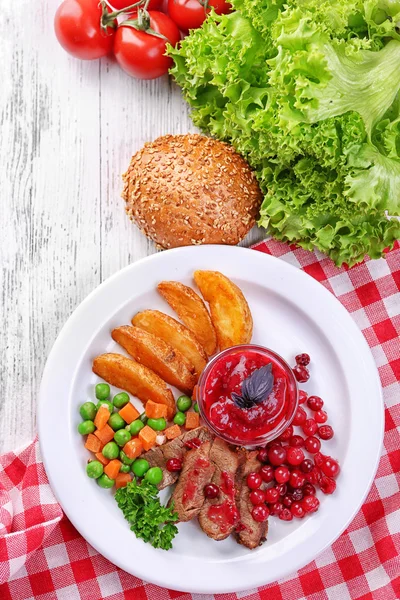 Carne di manzo con salsa di mirtilli rossi, fette di patate arrosto, verdure e panino sul piatto, su sfondo di legno a colori — Foto Stock