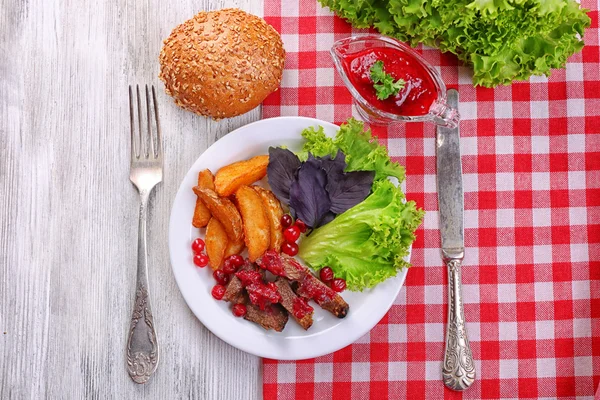Яловичина з журавлинним соусом, смажені скибочки картоплі, овочі та булочка на тарілці, на кольоровому дерев'яному фоні — стокове фото