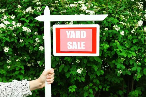 Деревянный двор Продажа знак в женской руке на зеленый куст и цветы фон — стоковое фото