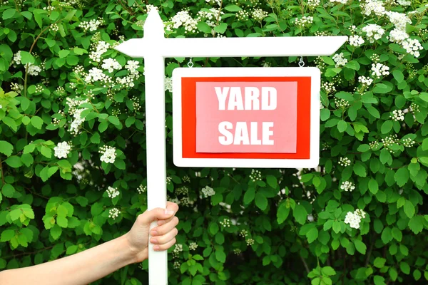 Sinal de venda de madeira Yard na mão feminina sobre arbusto verde e flores fundo — Fotografia de Stock