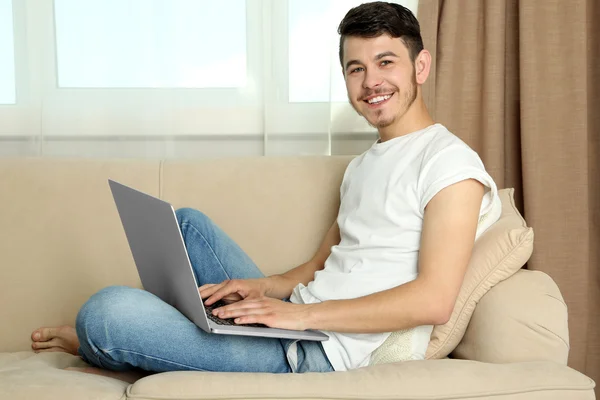Handsome young man sitting on sofa and using laptop in room — Stock Photo, Image