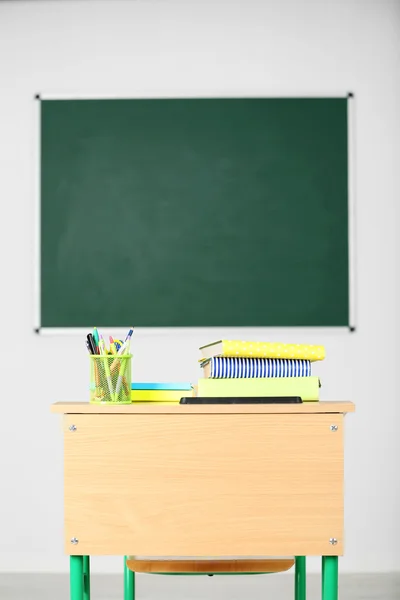 Wooden desk with stationery and chair in class on blackboard background — Stock Photo, Image