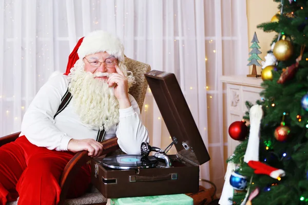 Weihnachtsmann sitzt in bequemem Stuhl in der Nähe von Retro-Plattenspieler zu Hause — Stockfoto