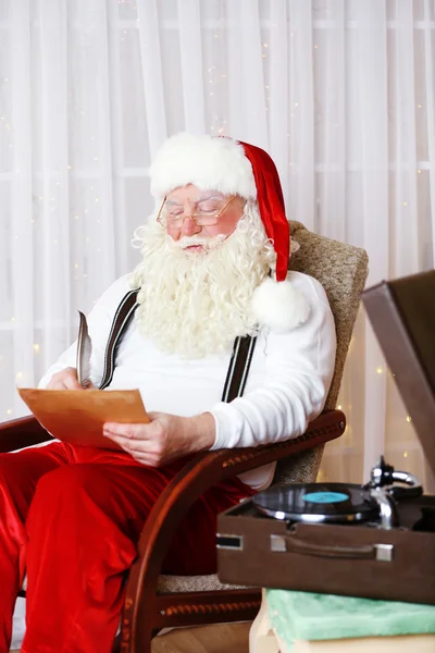 Santa Claus siedzi z listy życzeń dzieci w wygodnym fotelu w domu — Zdjęcie stockowe