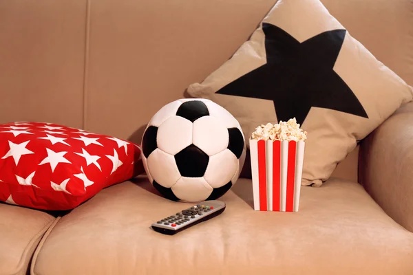 Soccer ball, remote control and box of popcorn on comfortable sofa, indoors — Stock Photo, Image