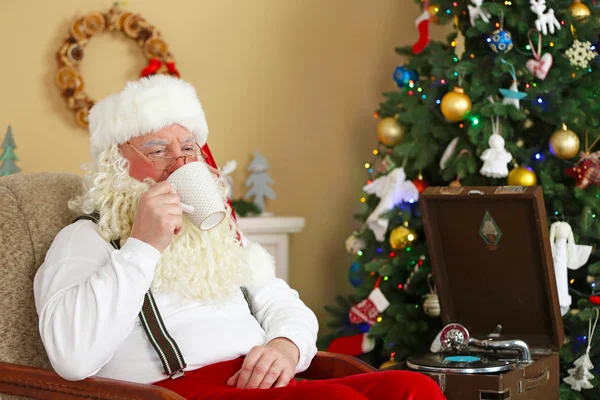 Santa claus zitten met kop warme dranken in de comfortabele stoel in de buurt van open haard thuis — Stockfoto