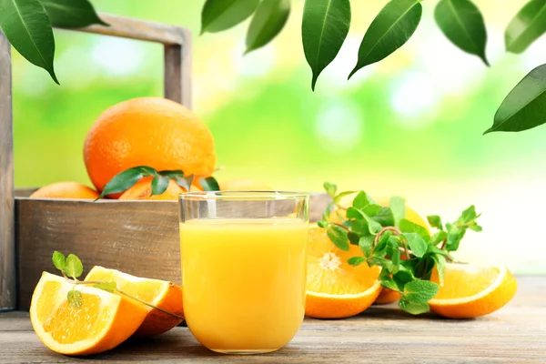 Glass of orange juice with oranges in crate on wooden table and natural background — Stock Photo, Image
