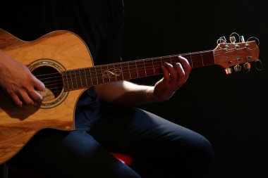 Genç adam akustik gitarla karanlık arka planda çalıyor.