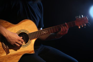 Genç adam akustik gitarla karanlık arka planda çalıyor.