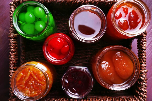 Homemade jars of fruits jam in wicker basket and color wooden background — Stock Photo, Image