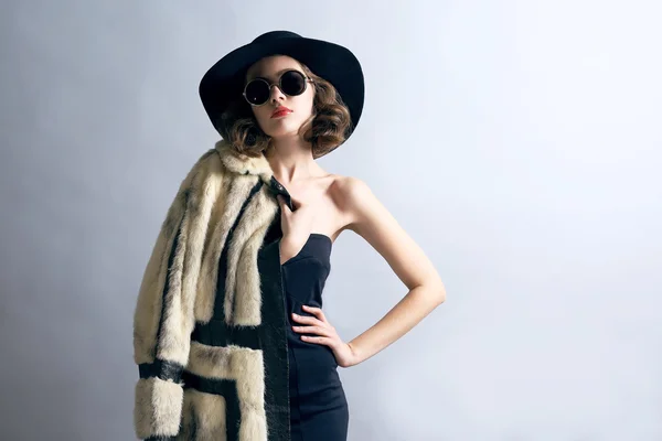 Retrato de modelo hermoso en abrigo de piel, sombrero y gafas de sol sobre fondo gris — Foto de Stock