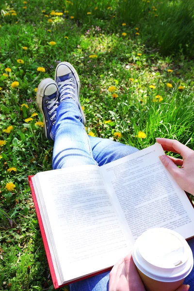 Молода жінка з книгою і чашкою кави сидить на зеленій траві на відкритому повітрі — стокове фото