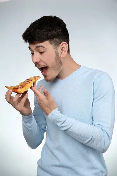 Junger Mann isst Stück Pizza auf grauem Hintergrund — Stockfoto