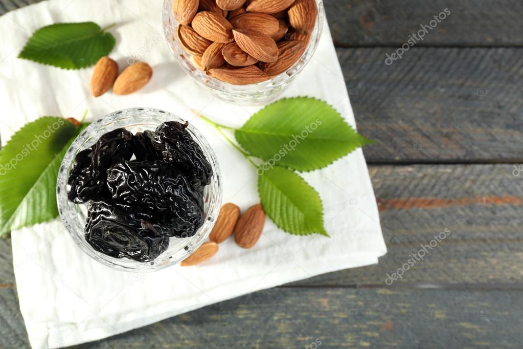 Prunes and almonds in glasses with leaves on wooden table, top view