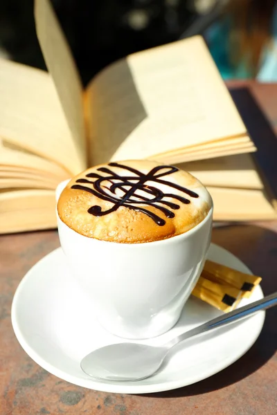 Tazas de capuchino con clave de agudos en espuma y libro en la mesa en la cafetería — Foto de Stock