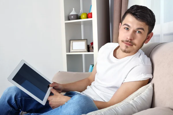 Hombre joven guapo sentado en el sofá y el uso de la tableta en la habitación —  Fotos de Stock