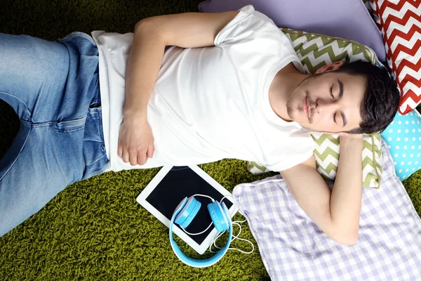 Красивый молодой человек в наушниках и планшете лежит на ковре в комнате — стоковое фото