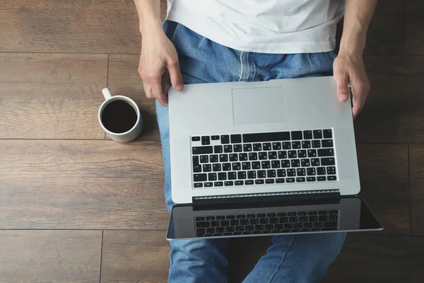 Young man sitting on floor with laptop and cup of coffee in room — Stock Photo, Image