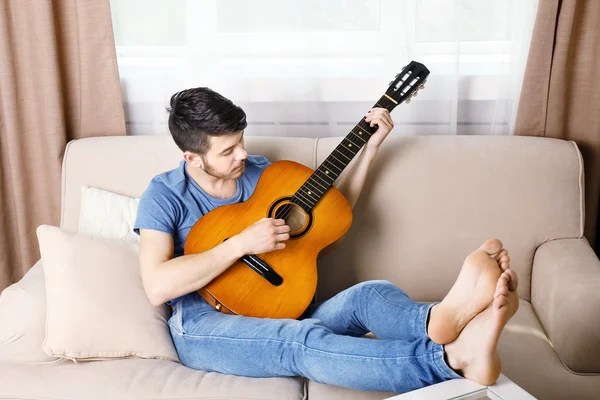 Młody człowiek z gitarą na kanapie w pokoju — Zdjęcie stockowe