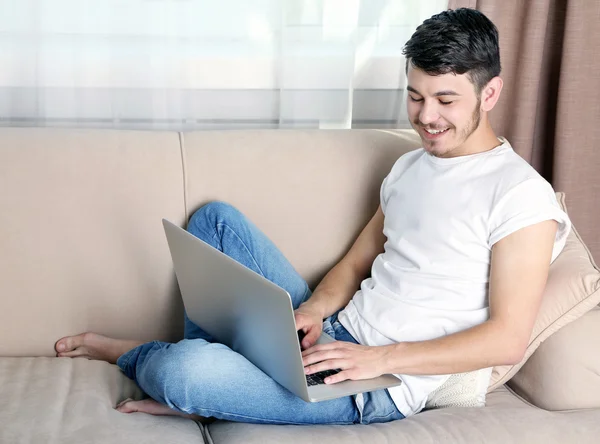 Pohledný mladý muž sedí na pohovce a používání notebooku v pokoji Stock Fotografie