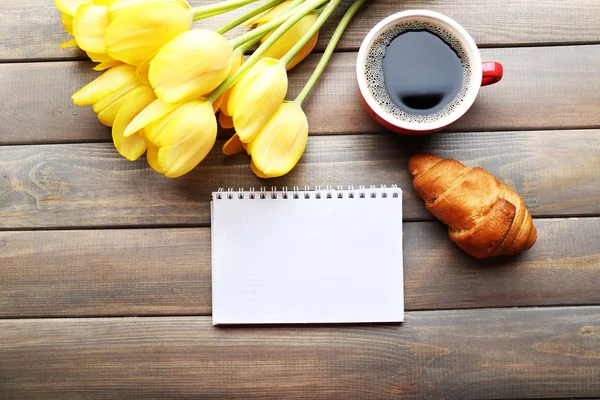 Xícara de café com croissant fresco, tulipas e folha de papel em branco sobre fundo de madeira — Fotografia de Stock