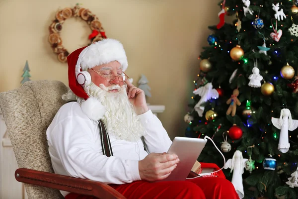 Santa Claus sentado con la tableta digital en silla cómoda cerca del árbol de Navidad en casa — Foto de Stock
