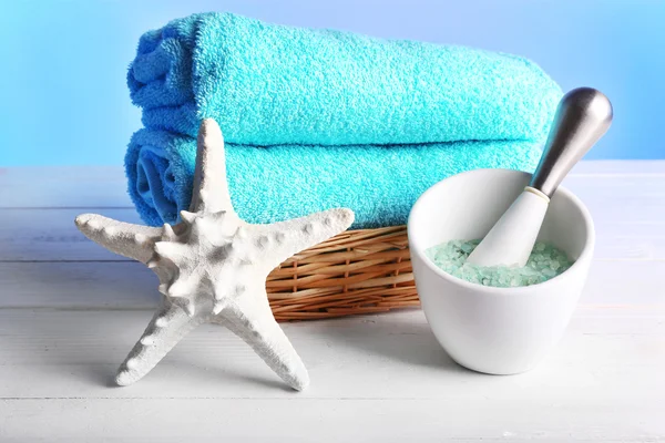 Warmgewalste handdoeken in rieten mand met zeester en zee zout op houten tafel en lichte kleurrijke achtergrond — Stockfoto