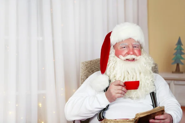 Weihnachtsmann sitzt mit Buch im bequemen Stuhl am heimischen Kamin — Stockfoto